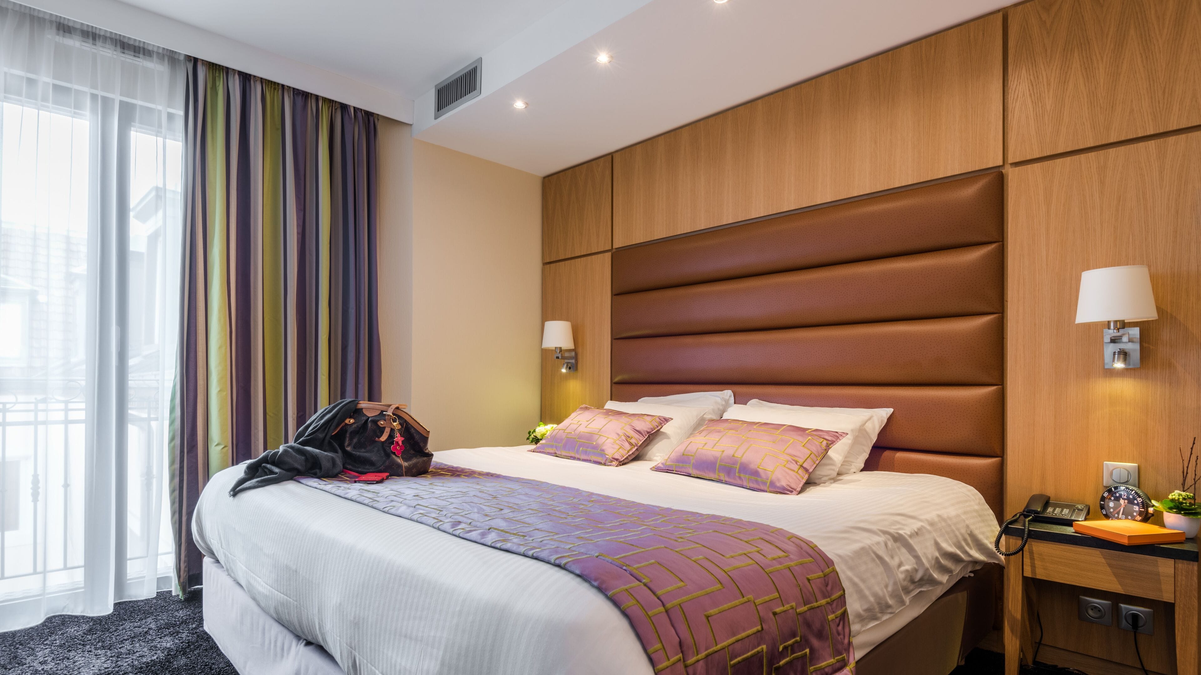 A hotel room with a large bed, a nightstand, a lamp, and a view of the outside through sheer curtains.