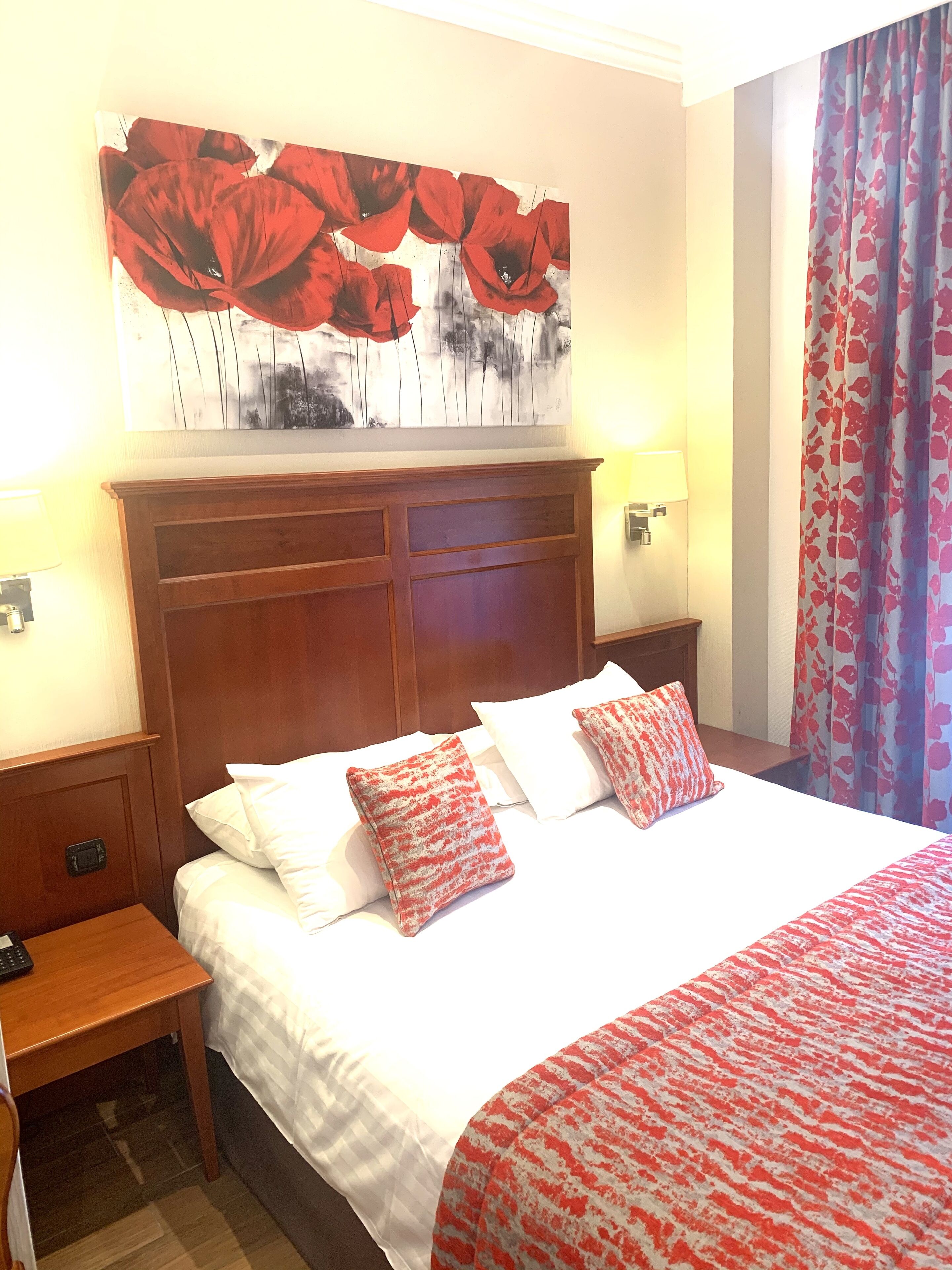 A hotel room with a large bed, two bedside tables, a wooden headboard, a painting above the bed, and a window with curtains.