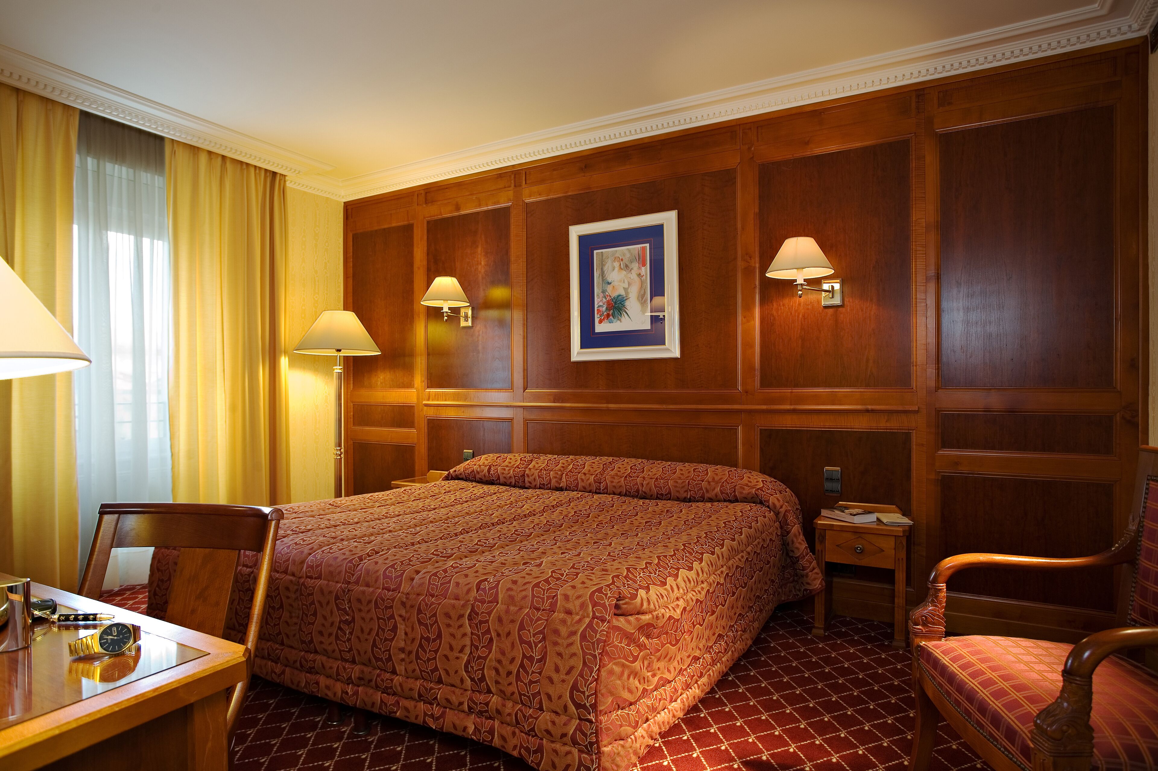A hotel room with a large bed, two bedside lamps, a wooden headboard, a small table with a clock, and a chair.