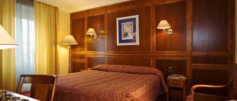 A hotel room with a large bed, two bedside lamps, a wooden headboard, a small table with a clock, and a chair.