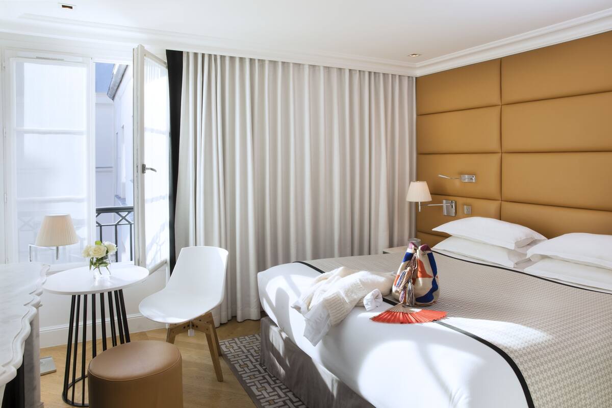 A hotel room with a bed, a small table with a lamp, a chair, and a window with curtains.