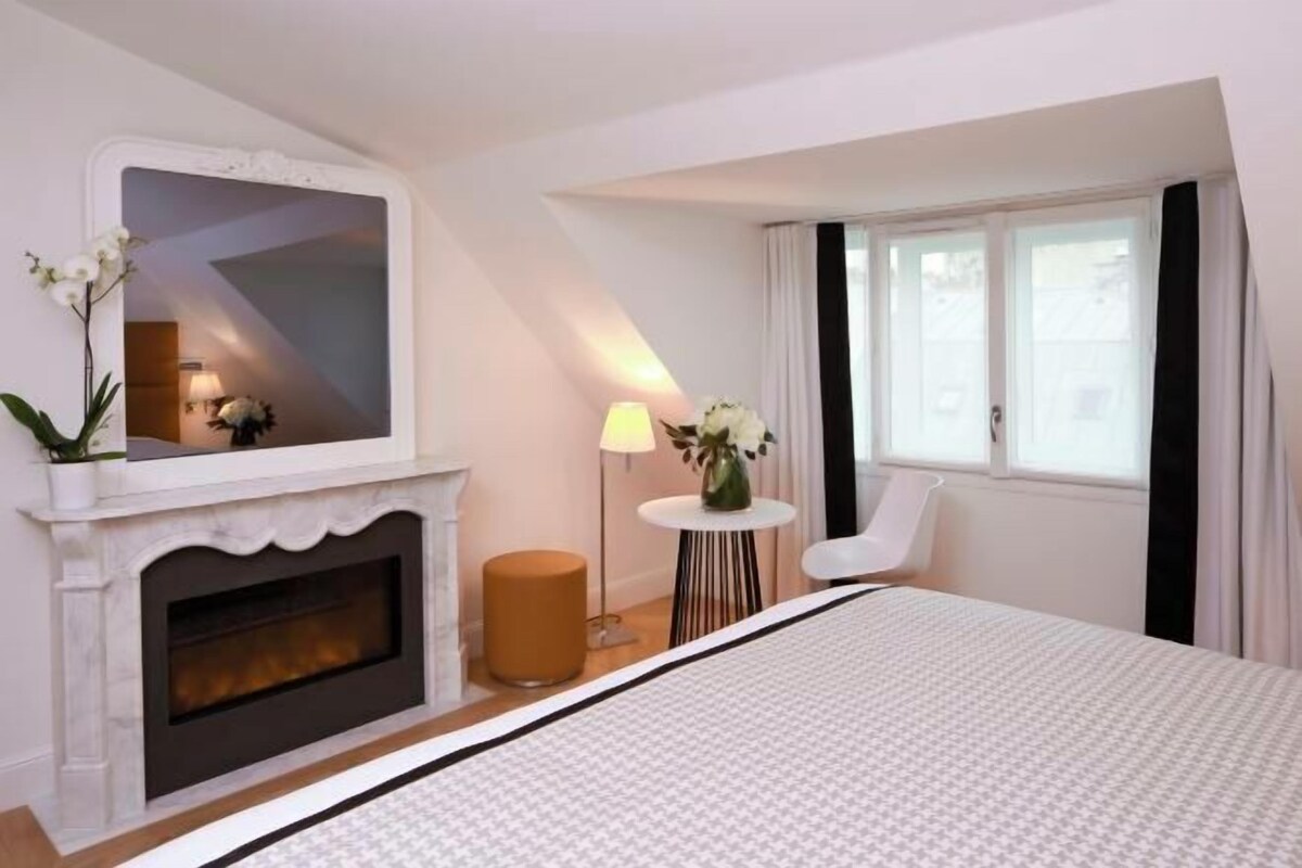 A hotel room with a fireplace, a bed, a mirror, a small table with a lamp, and a chair.