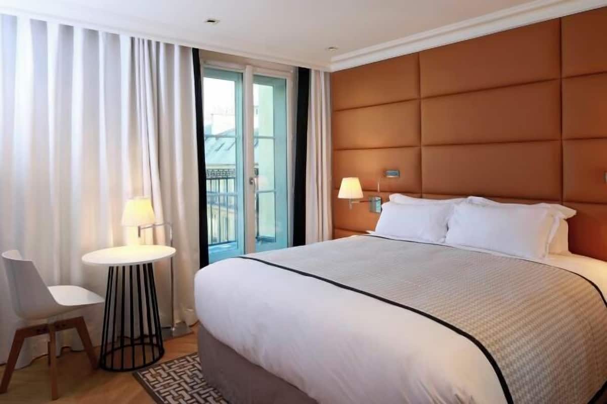 A hotel room with a large bed, a small round table, a chair, a balcony with curtains, and a headboard.