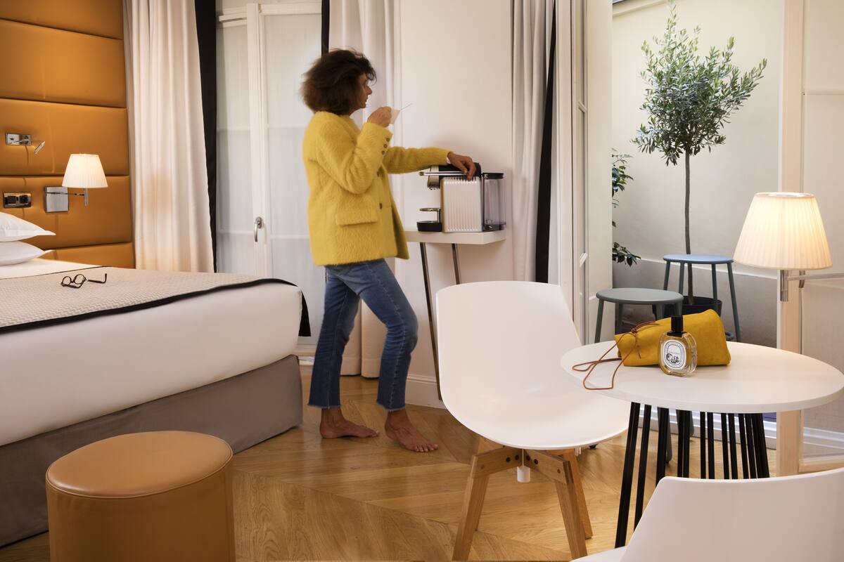 A hotel room with a bed, a bedside table with a lamp, a small table with a chair, and a potted plant.