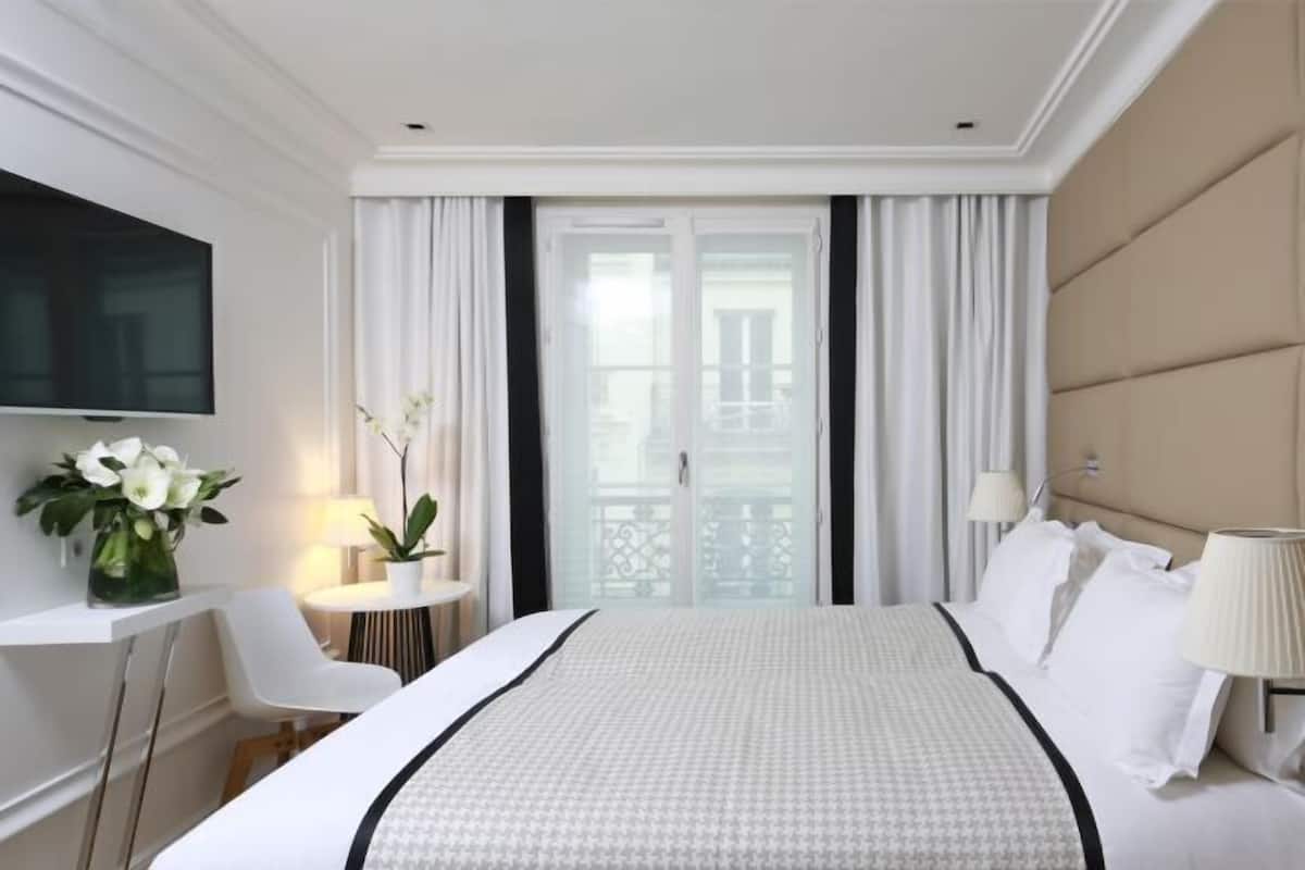 A hotel room with a bed, a TV, a desk with a lamp, a chair, and a view of the outside through a glass door.