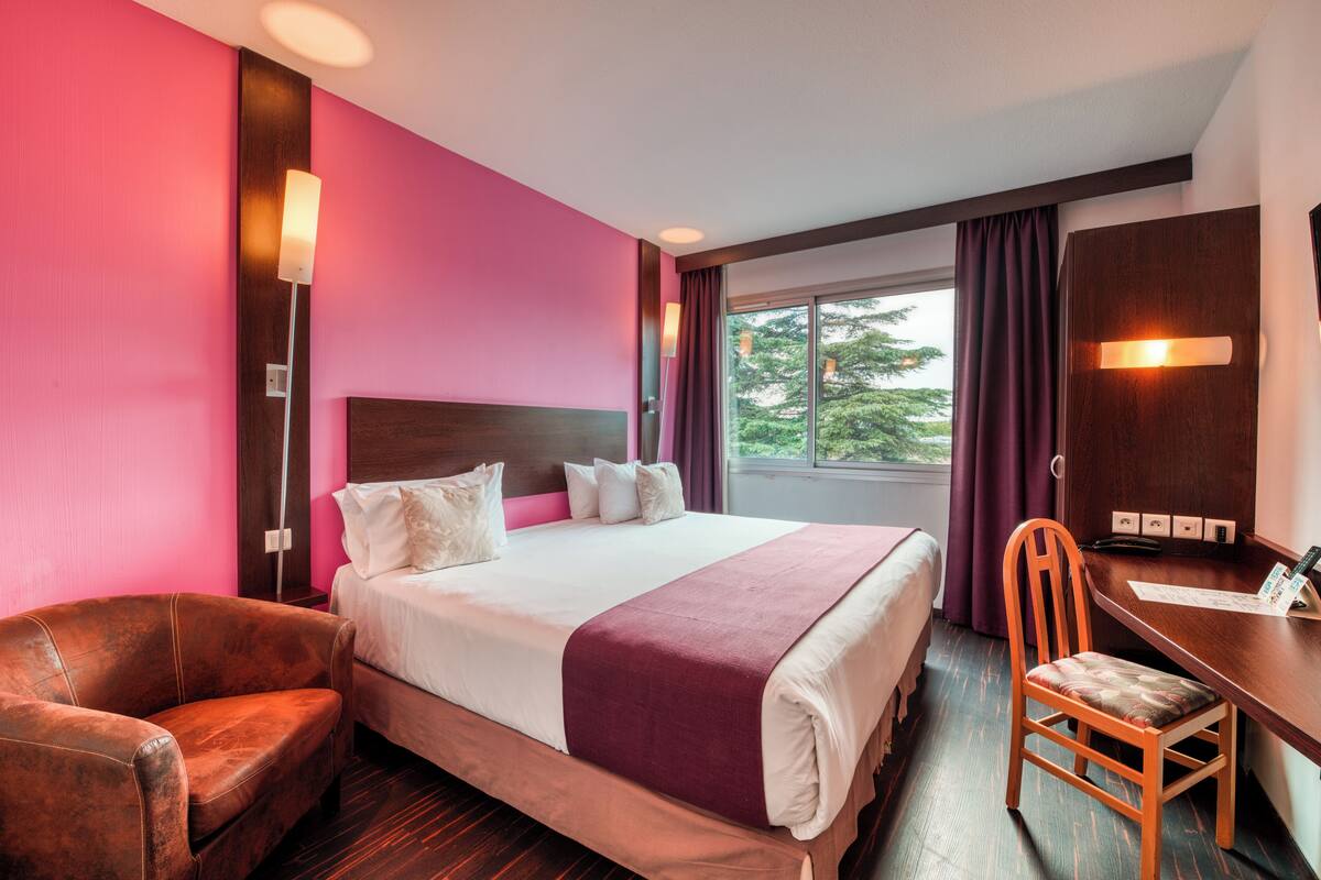 Habitación de hotel con una cama grande, un escritorio, una silla y vistas a árboles a través de la ventana.
