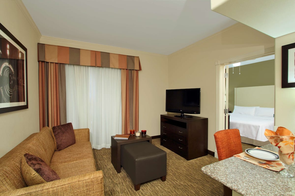 Habitación de hotel con sofá, una mesita con un vaso, televisión, cama y puerta que da a otra habitación.