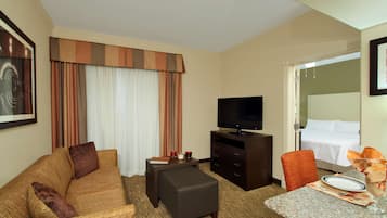 Habitación de hotel con sofá, una mesita con un vaso, televisión, cama y puerta que da a otra habitación.