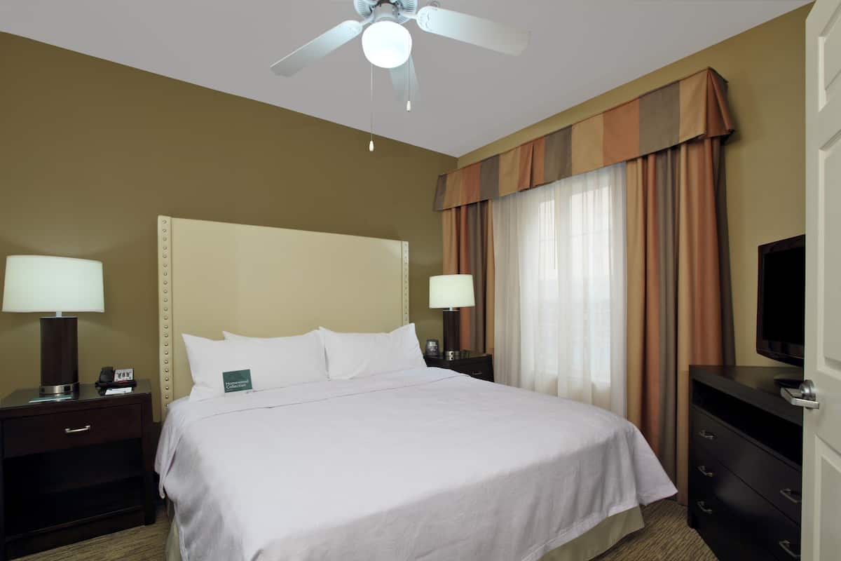 Habitación de hotel con cama, dos mesitas de noche con lámparas, televisión, ventilador de techo y ventana con cortinas.