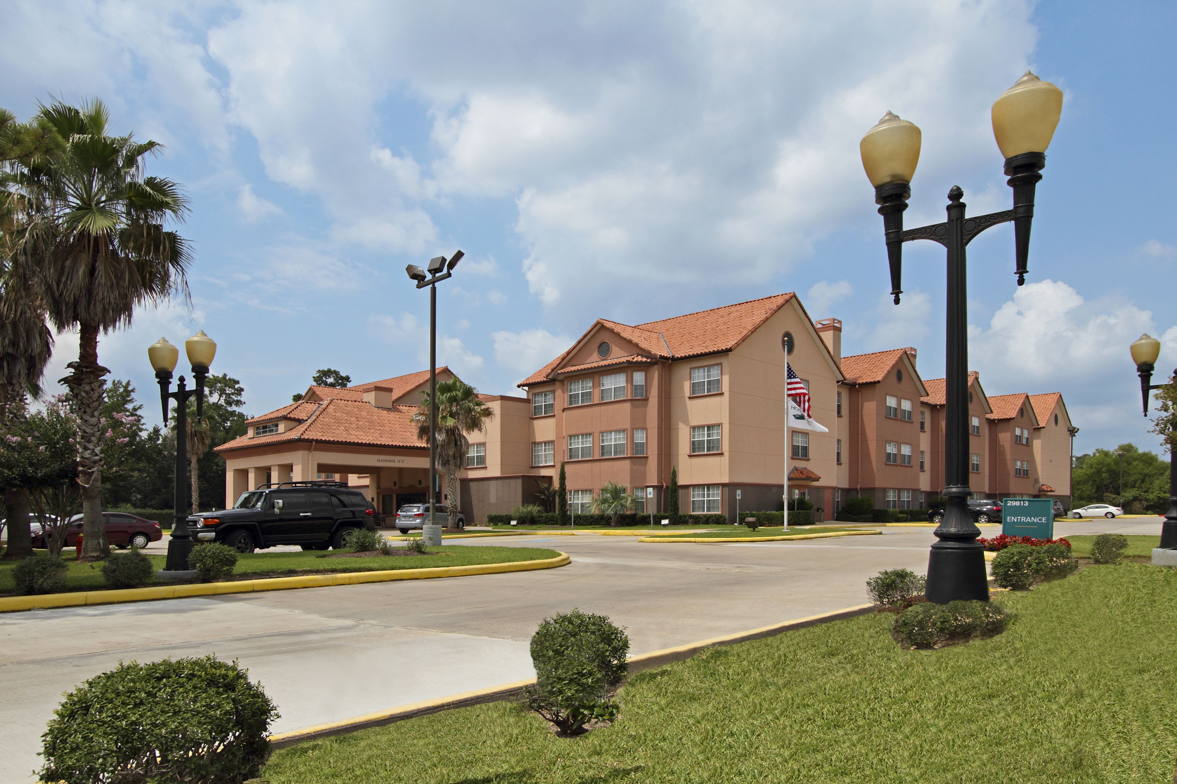 A hotel with a red-tiled roof, multiple windows, and a flag. There are cars parked in front, and a well-maintained lawn with small bushes.