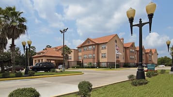 A hotel with a red-tiled roof, multiple windows, and a flag. There are cars parked in front, and a well-maintained lawn with small bushes.