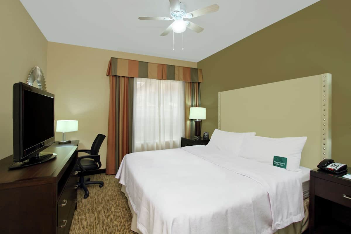 Una habitación de hotel con una cama, un escritorio con televisor, una silla, una lámpara y un ventilador de techo.