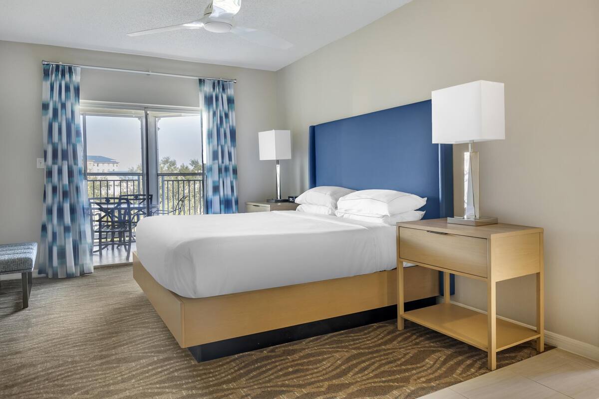Una habitación de hotel con cama, lámparas de noche, mesita de noche, una silla y vistas al exterior.