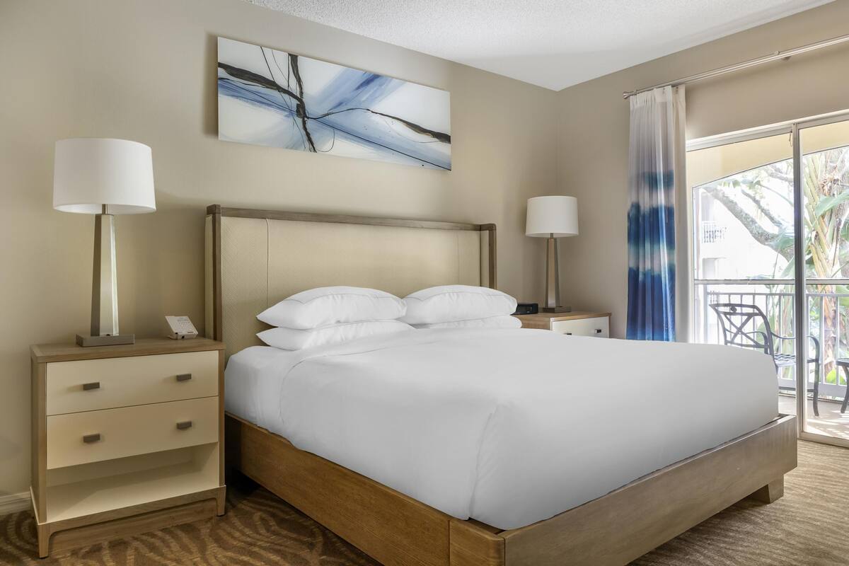 Habitación de hotel con cama, dos mesitas de noche, un cuadro y vistas al exterior.