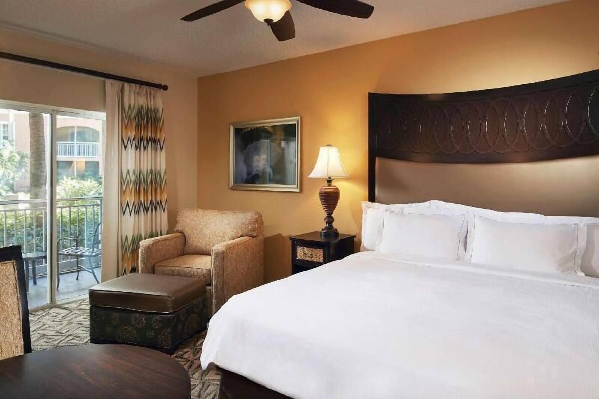 Habitación de hotel con cama, una silla, una mesa, lámpara, un cuadro y balcón con cortinas.