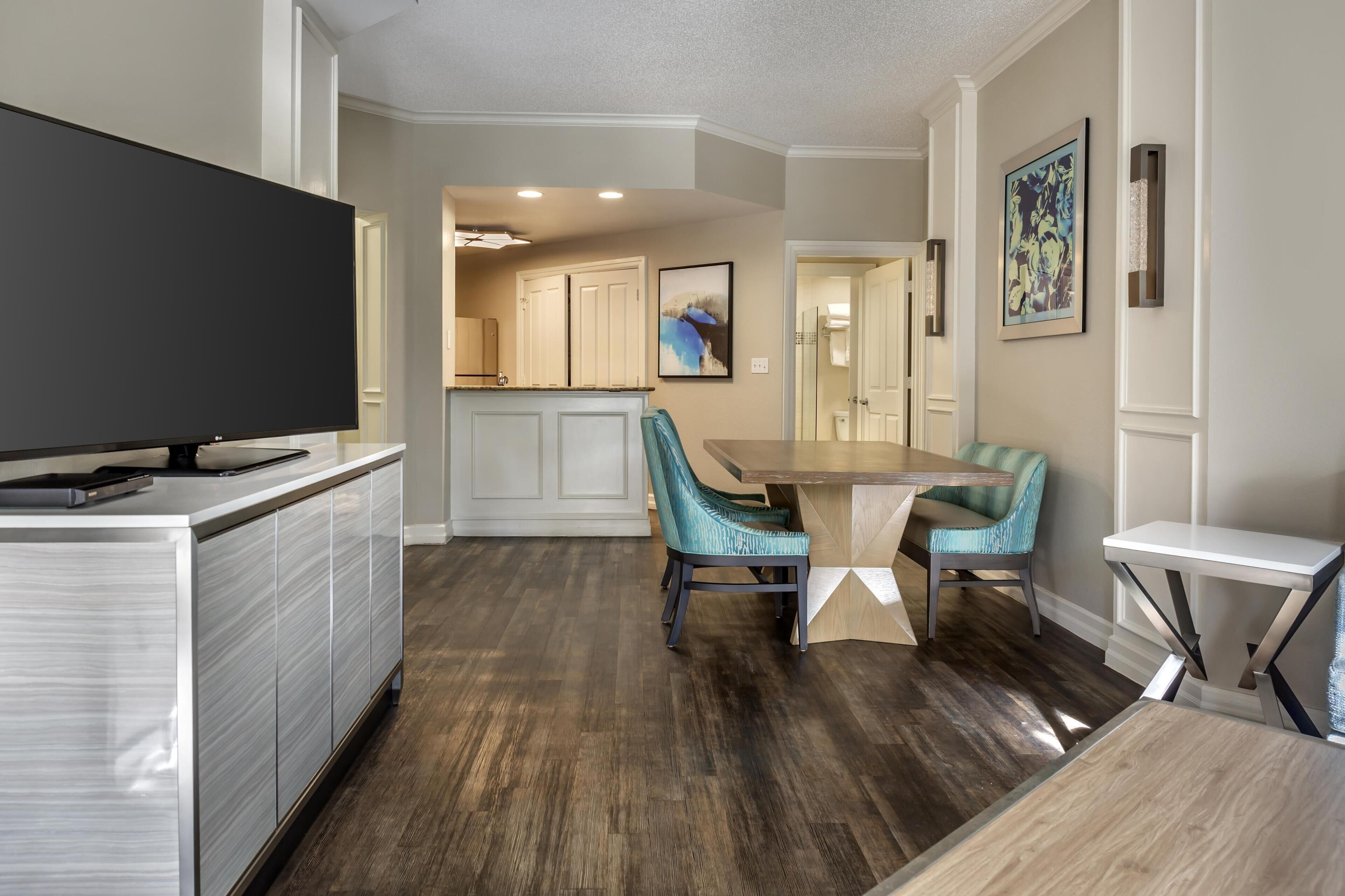 A modern kitchen with a flat-screen TV, wooden cabinets, and a dining area with teal chairs and a wooden table.
