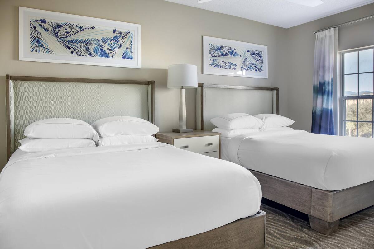 Una habitación de hotel con dos camas, una mesita de noche con lámpara y dos cuadros enmarcados en la pared.