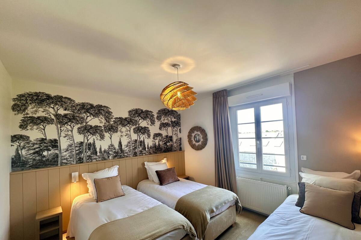 A hotel room with two beds, a large window with curtains, a wall mural of trees, a hanging light fixture, and a radiator.