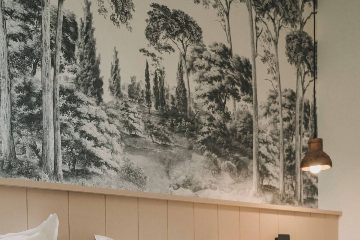 A bedroom with a bed, pillows, a wall-mounted lamp, and a forest-themed mural.