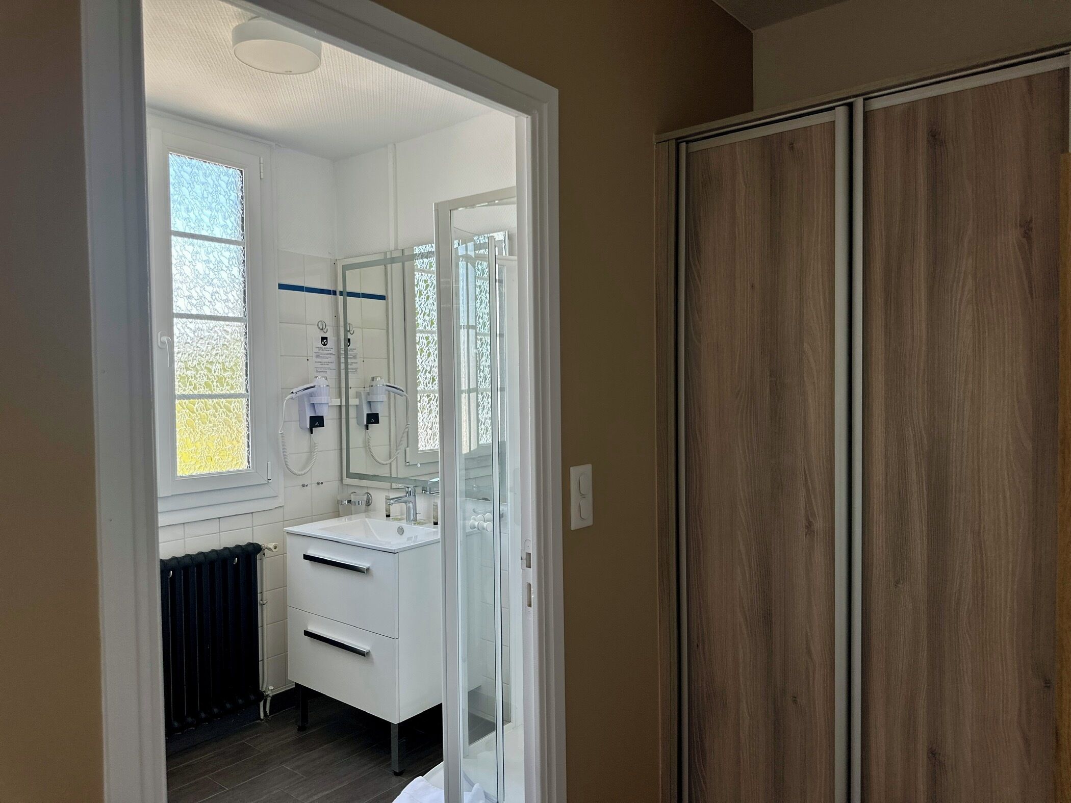 A modern bathroom with a white sink, mirror, and toilet. There is a window with a decorative screen, and a radiator is visible.