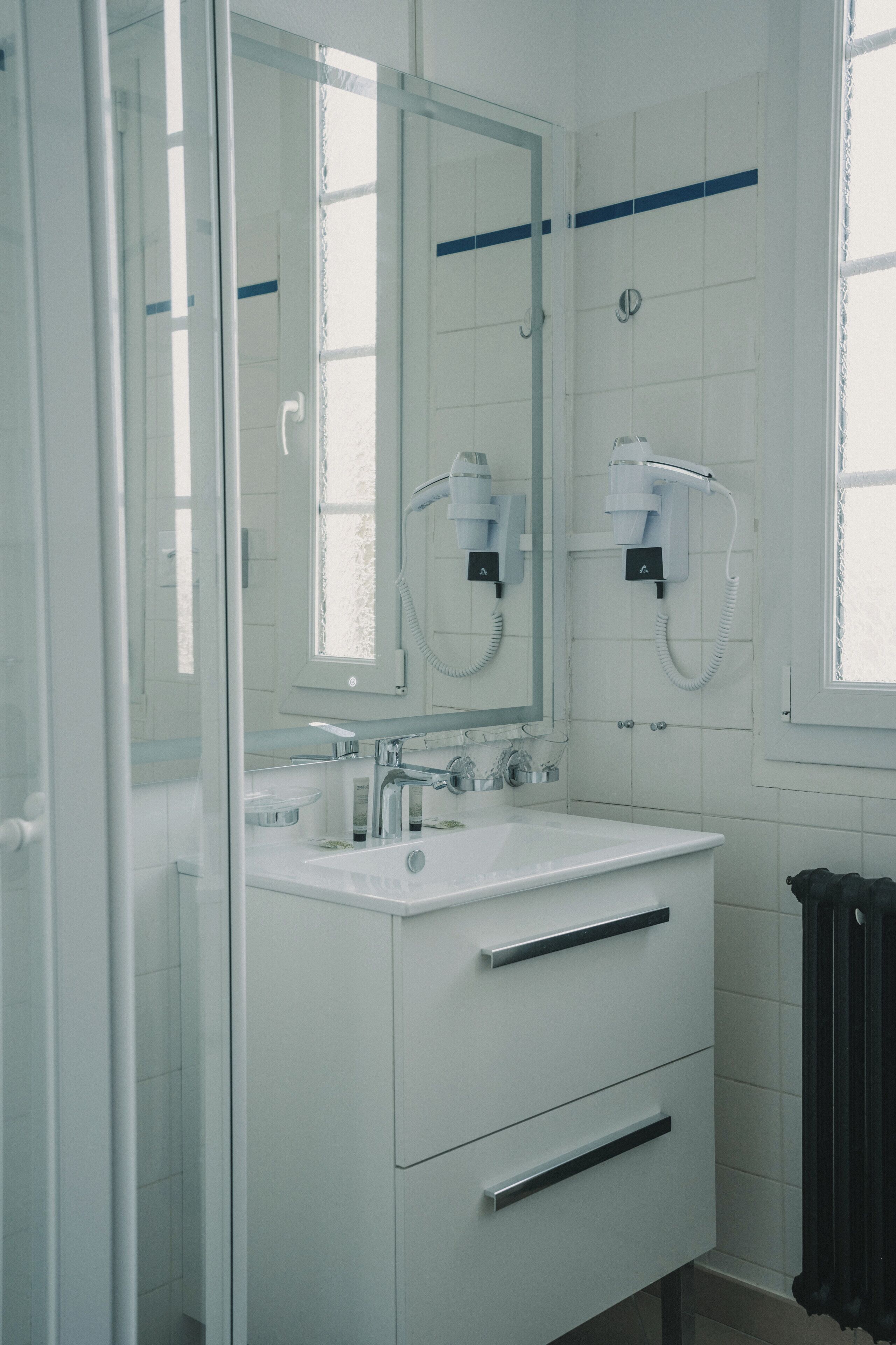 A modern bathroom with a glass shower enclosure, a white sink with a mirror above it, and a hairdryer mounted on the wall.