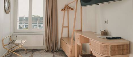 A room with a wooden desk, a television mounted on the wall, a radiator, a window with curtains, and a chair.
