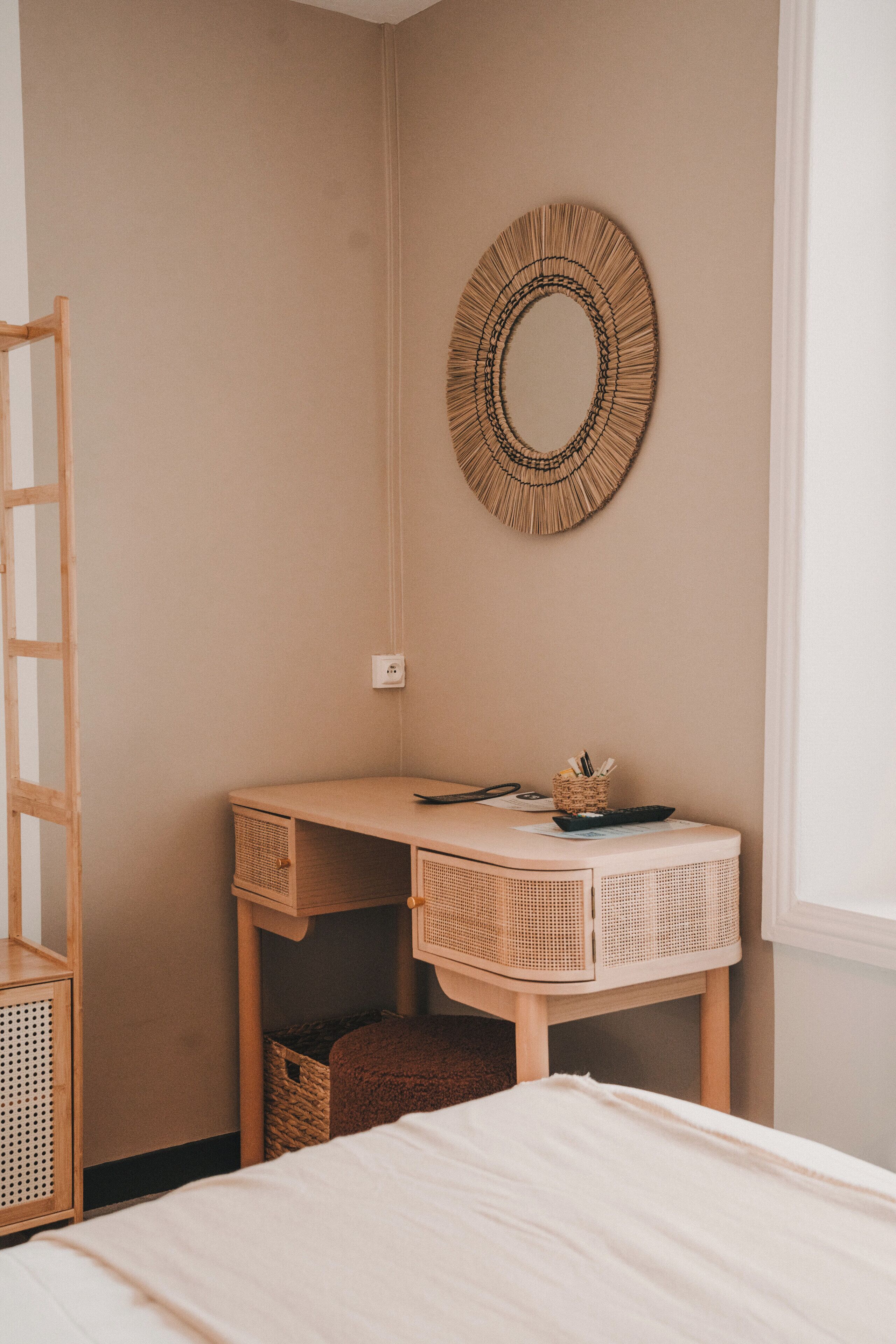 A bedroom with a bed, a wooden desk, a wicker wall hanging, and a window.