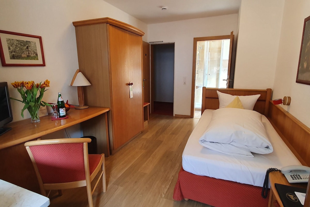 Une chambre d’hôtel avec un lit, un bureau équipé d’un ordinateur, une chaise, une armoire et une salle de bain visible à travers une porte ouverte.