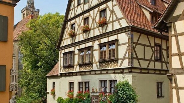 Un bâtiment traditionnel à colombages, avec une rue pavée, le clocher d’une église visible au loin et un arbre devant le bâtiment.