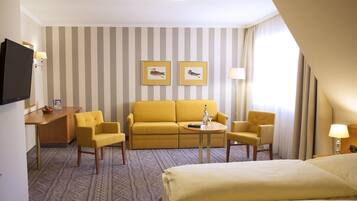 Ein Hotelzimmer mit gestreifter Wand, einem gelben Sofa, zwei Sesseln, einem kleinen Tisch mit Flasche und Glas, einem Fernseher und einem Bett.