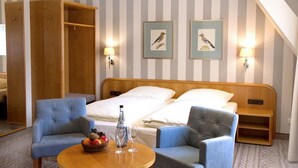 Une chambre d’hôtel avec un lit, deux fauteuils, une petite table avec des fruits et des verres, et deux tableaux encadrés au mur.
