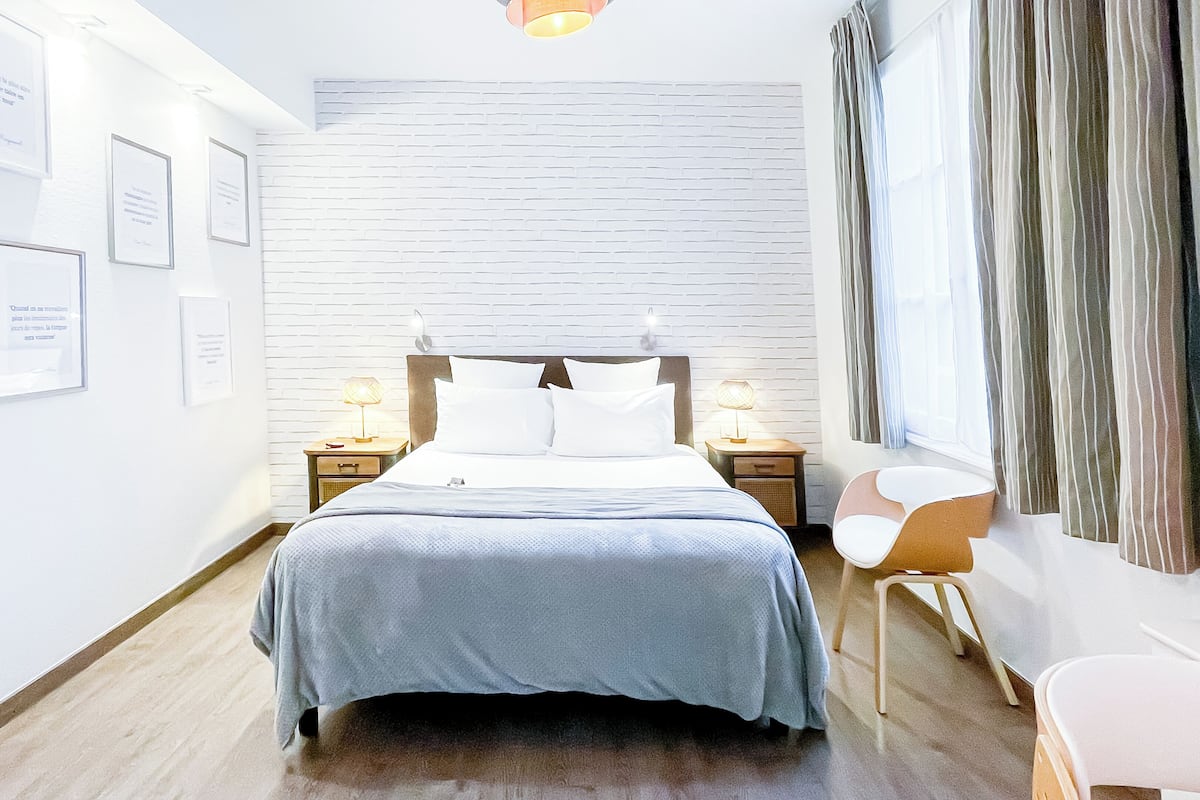 A hotel room with a bed, two bedside tables with lamps, a chair, a white brick wall, and framed certificates on the wall.