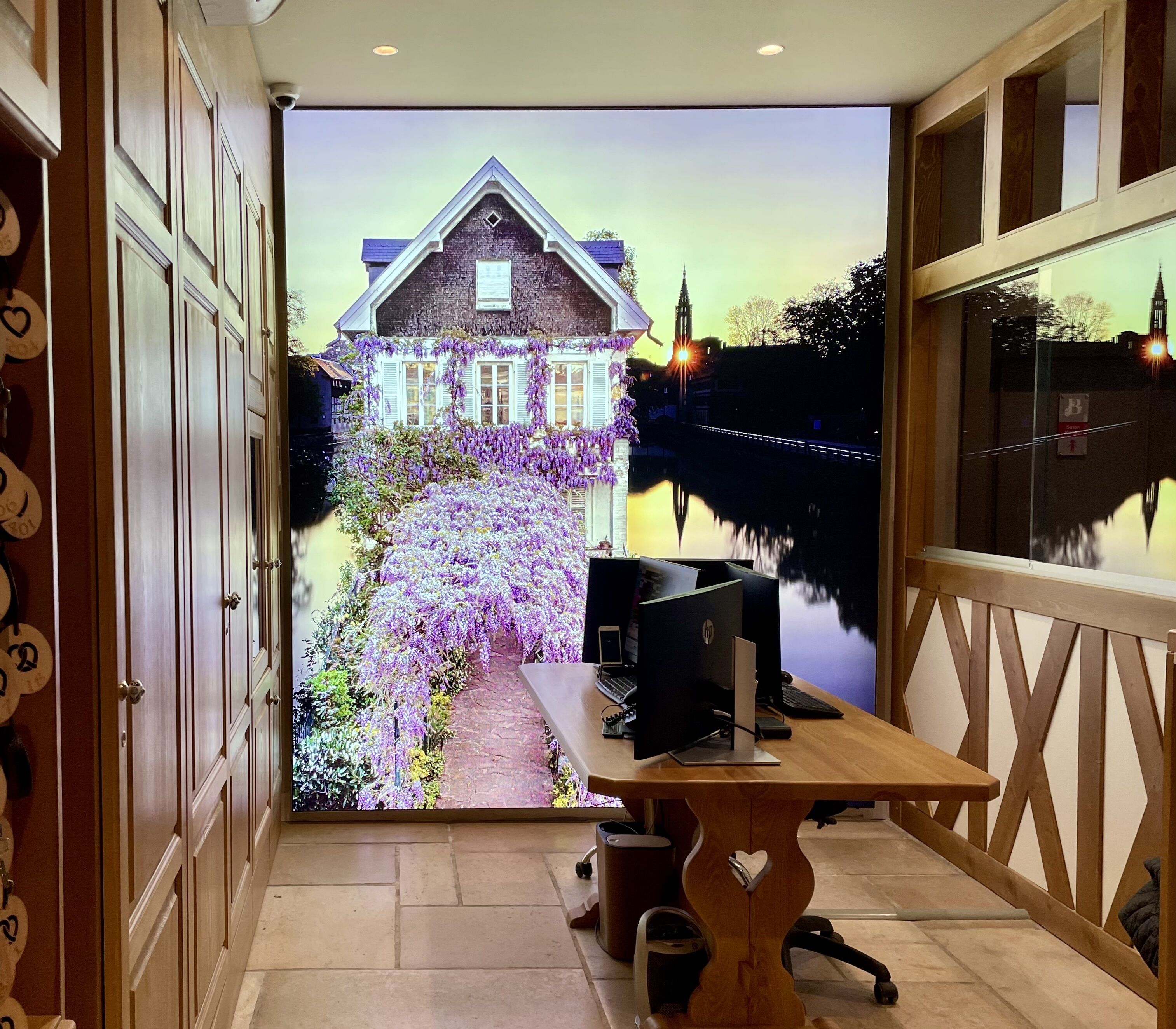 A room with a desk, computer, and a view of a house with a garden and a river.