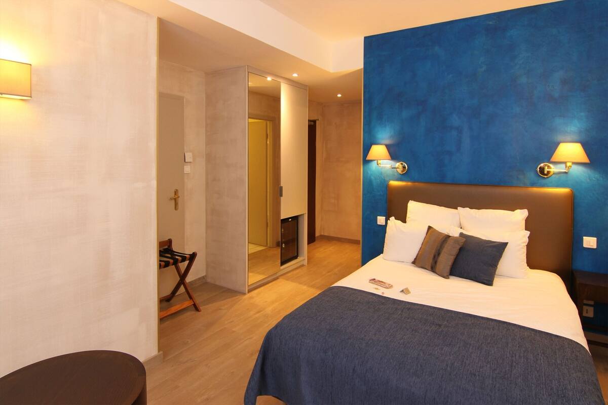 A hotel room with a bed, two wall-mounted lamps, a wooden floor, and a door leading to a bathroom.