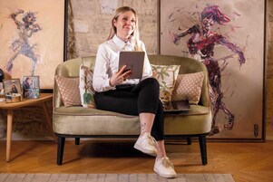 Eine Person sitzt auf einem grünen Sofa mit gemusterten Kissen und hält ein Tablet.