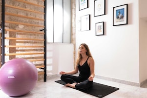A person meditating on a black mat in a room with a brick wall, a wooden rack, and framed pictures on the wall.