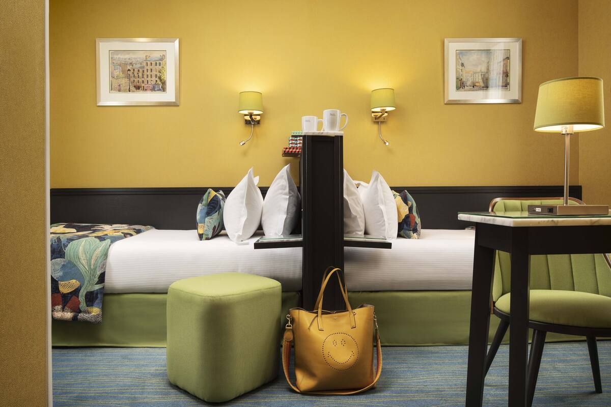 A hotel room with a bed, green ottoman, a table with a chair, a lamp, and two wall-mounted lamps.