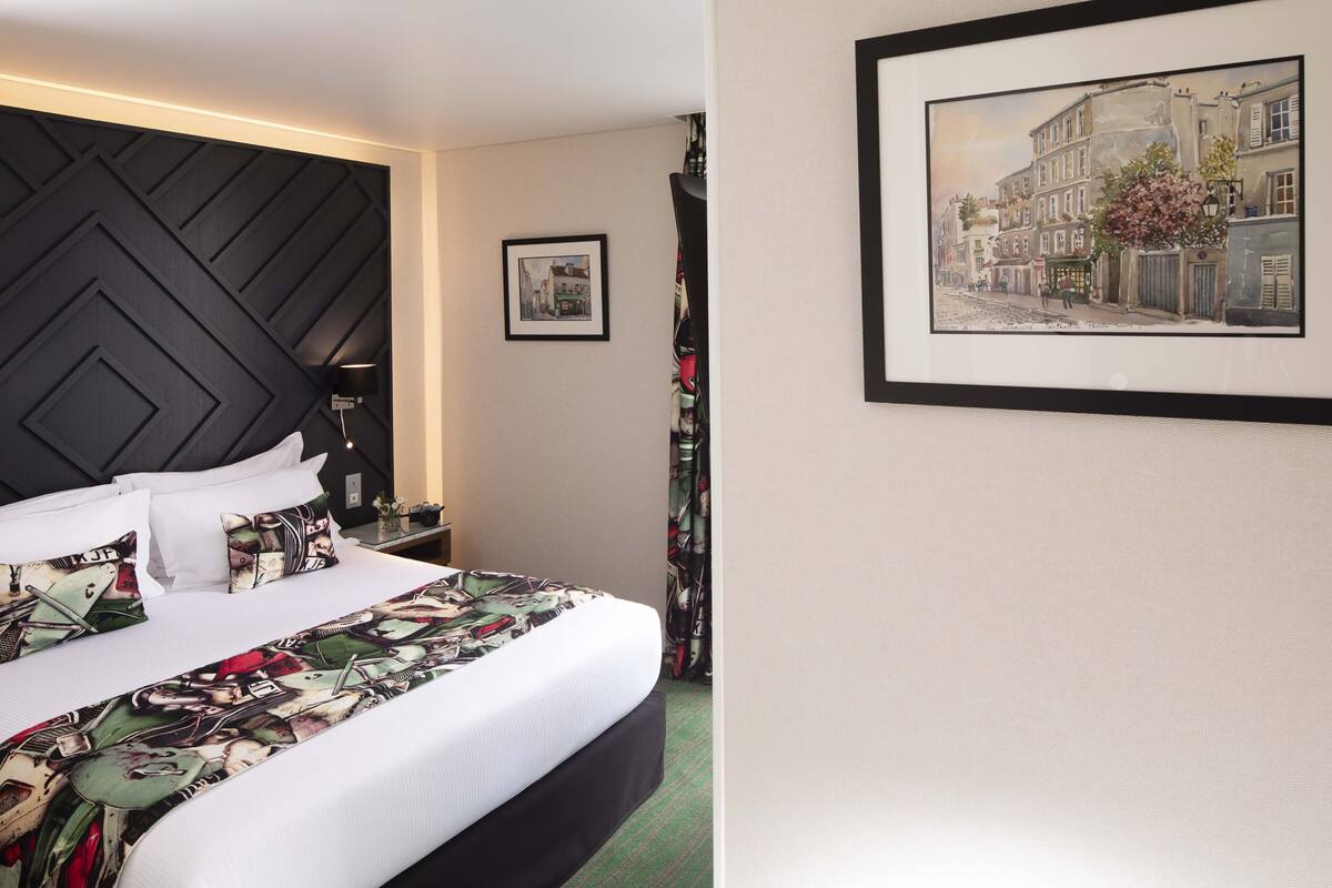 A hotel room with a large bed, a headboard with a geometric pattern, a framed picture on the wall, and a small table with a lamp.