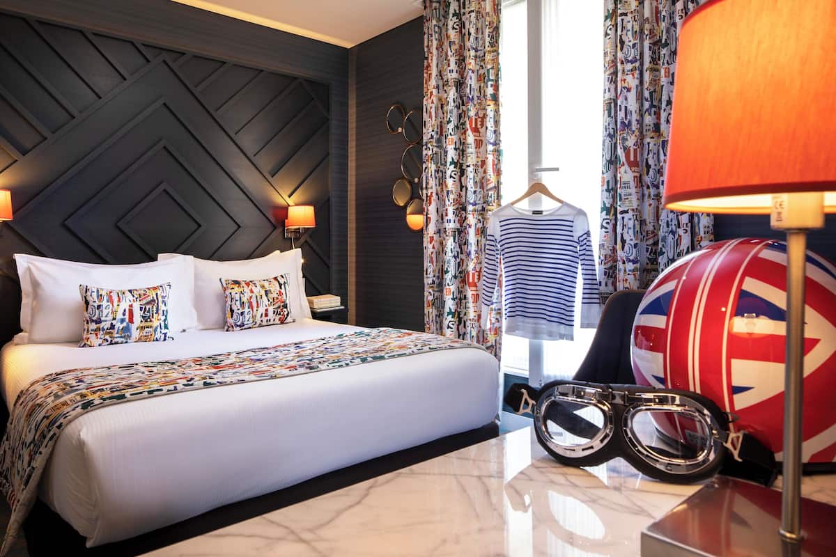 A hotel room with a large bed, a headboard with a geometric pattern, patterned curtains, a lamp, a clothing rack with a striped shirt, and a drum with a Union Jack design.