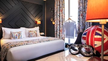 Habitación de hotel con una cama grande, cabecera con diseño geométrico, cortinas estampadas, una lámpara, un perchero con una camisa a rayas y un tambor con diseño de la Union Jack.