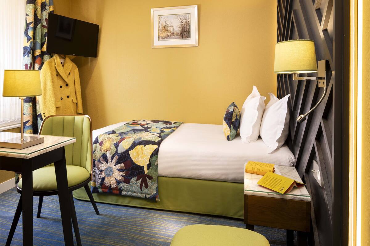A hotel room with a bed, a desk, a chair, a TV mounted on the wall, and a bedside table with a lamp.