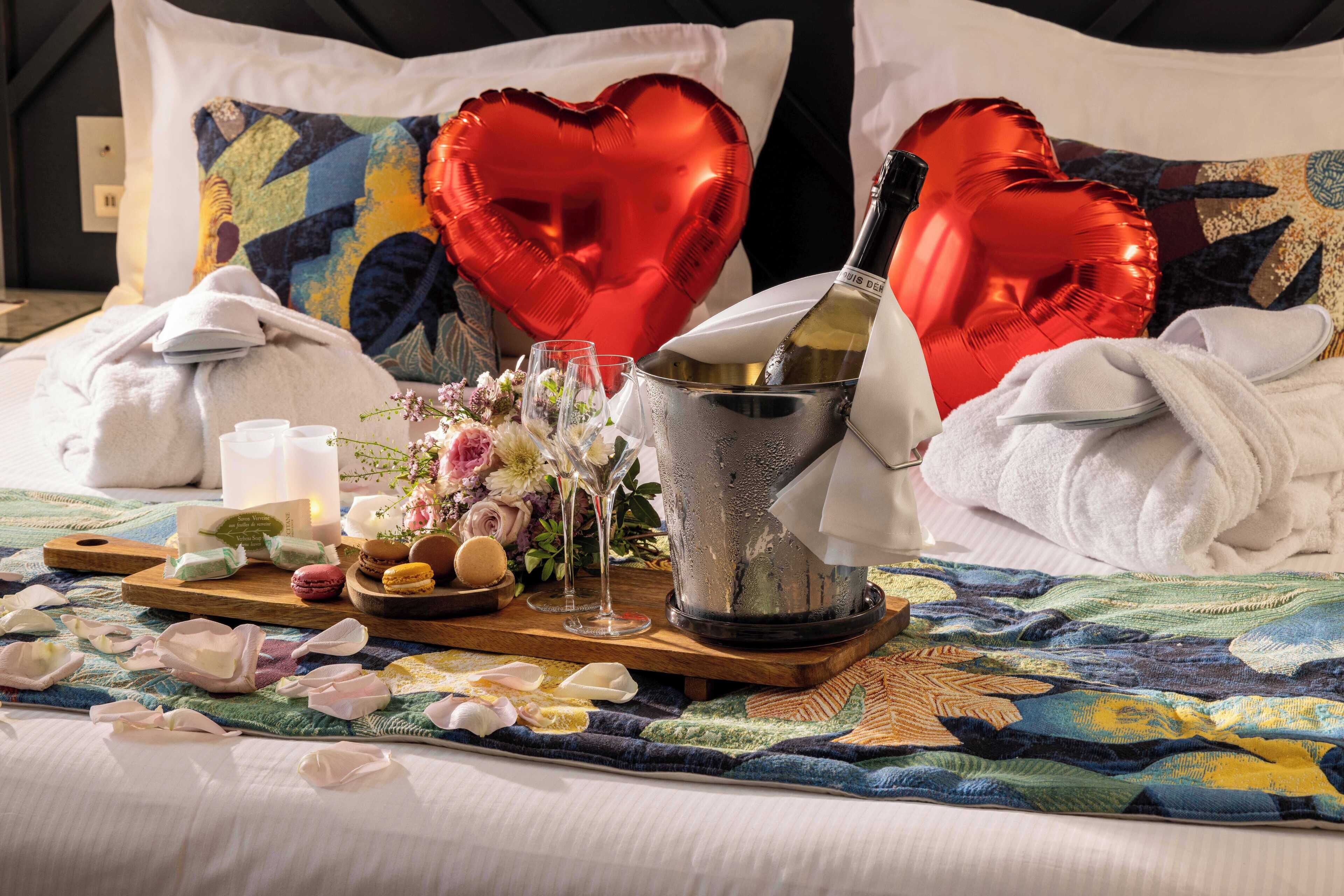 A bed with heart-shaped balloons, a champagne bottle, glasses, and a tray with snacks and flowers.