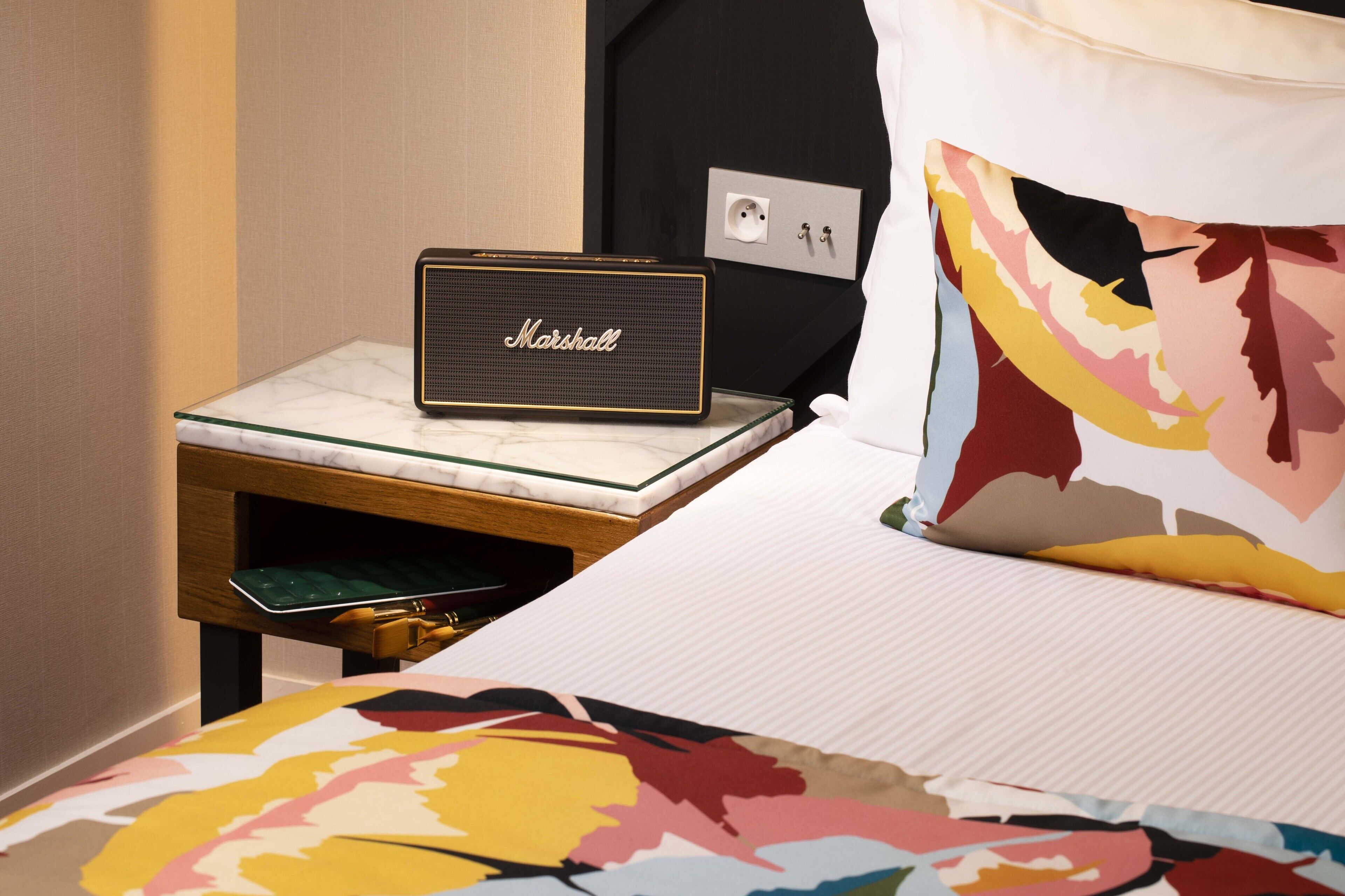 A hotel room with a bed, a Marshall speaker on a glass table, and a pillow with a colorful pattern.