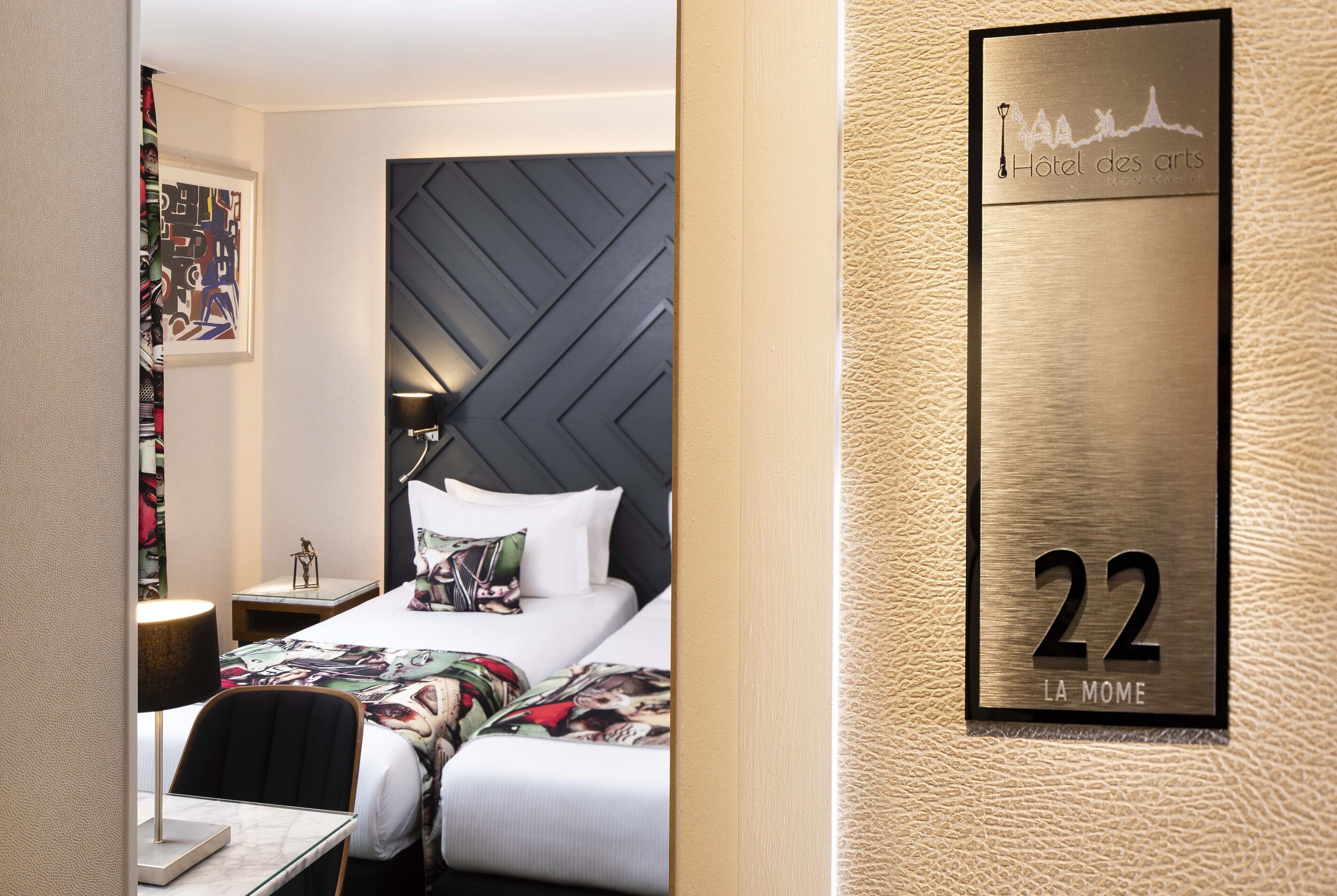 A hotel room with a modern design, featuring a large bed with a geometric headboard, a bedside table with a lamp, and a framed artwork on the wall. The room number is 22, and the hotel name is Hotel des Arts.