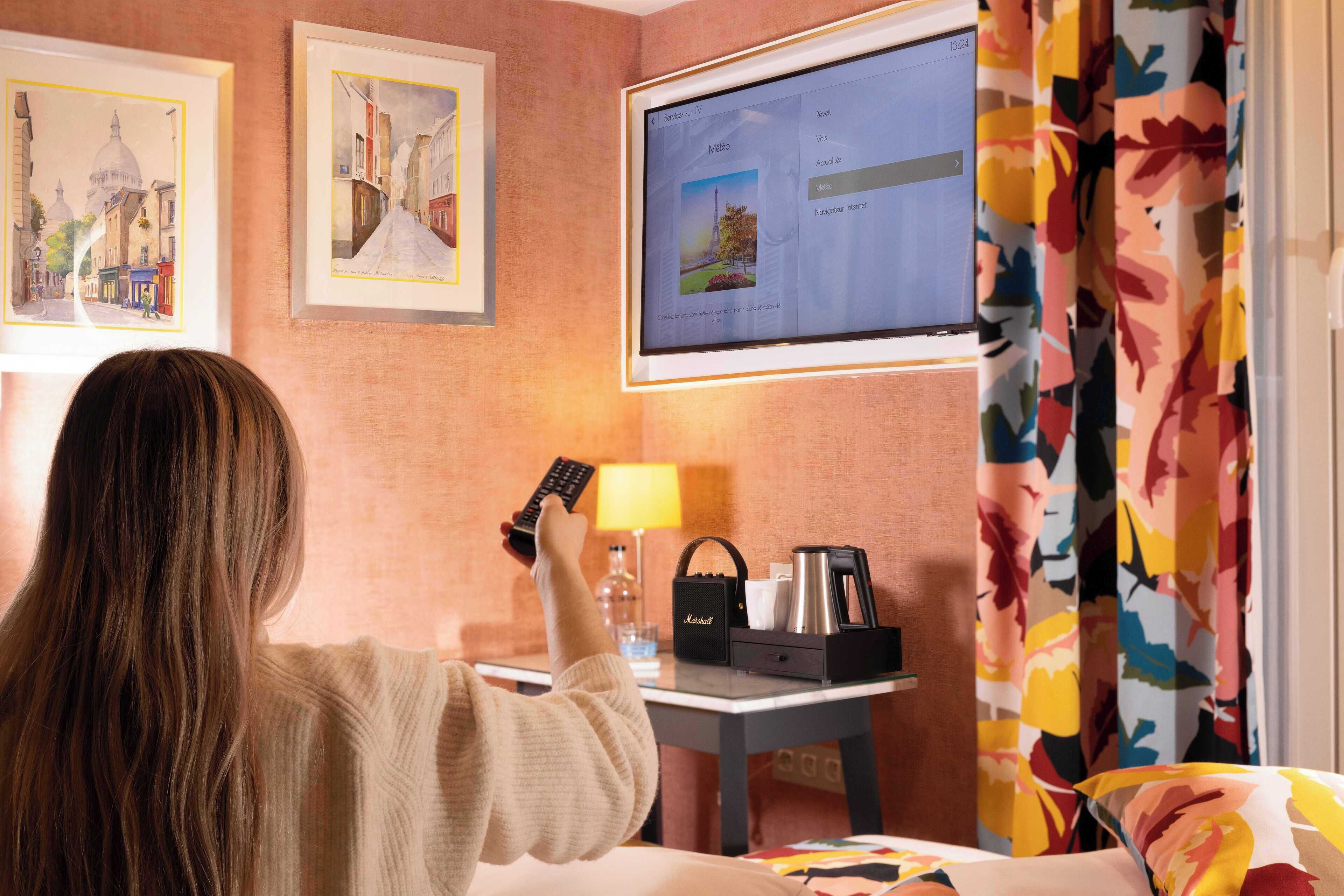 A hotel room with a bed, a desk with a laptop, a remote control, a coffee maker, and a small table with a glass and a bottle.