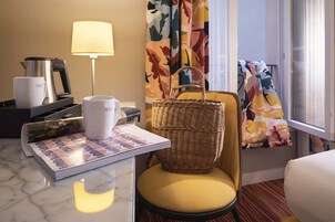 Habitación de hotel con cama, una silla con cojín amarillo, una cesta de mimbre, una bandeja con tazas y cafetera, una lámpara y una ventana con cortinas.