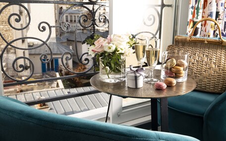 A balcony with a table set for two, featuring a vase of flowers, champagne glasses, and a basket of macarons.