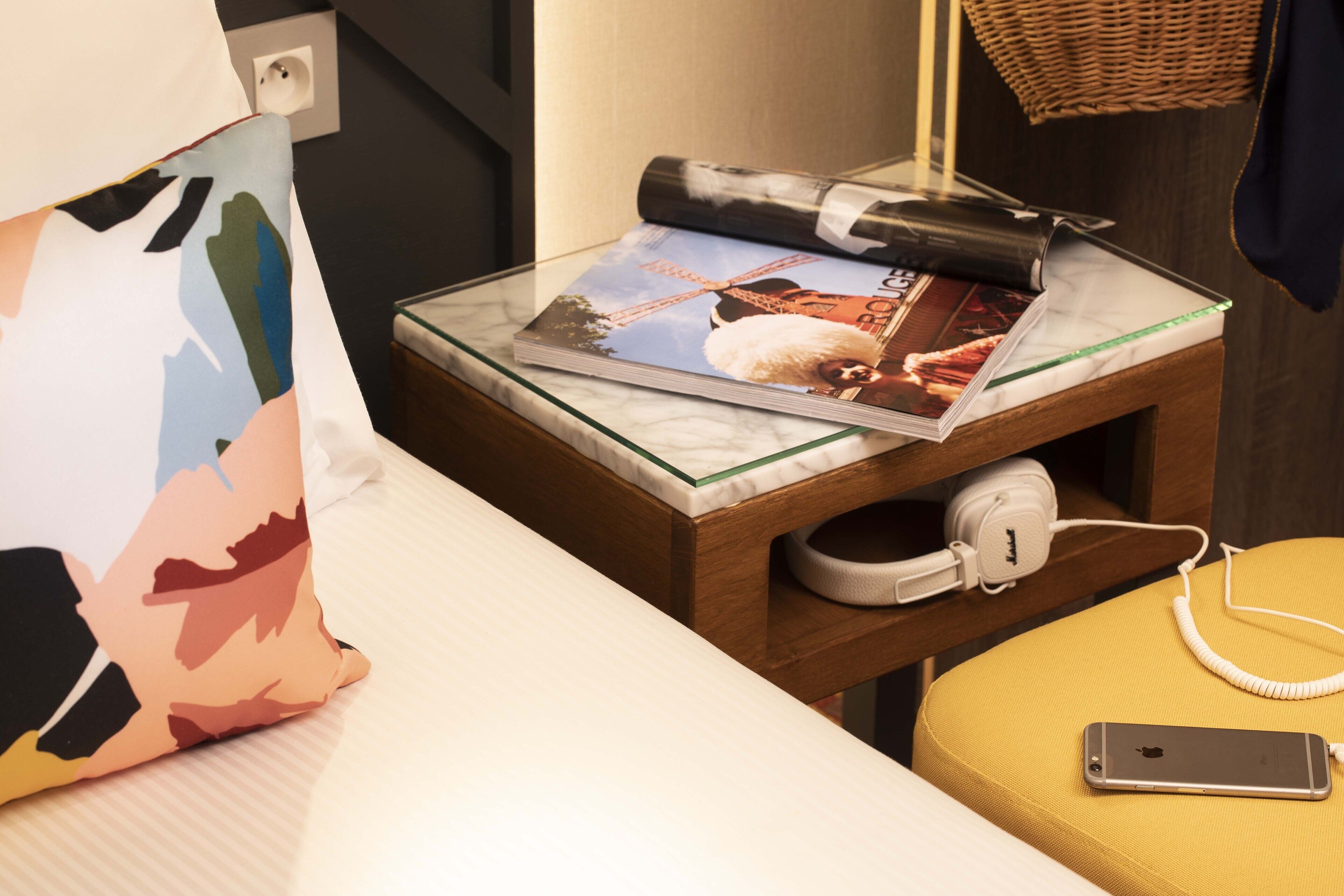 A neatly arranged hotel room with a bed, a small wooden side table with a magazine, a white charging station, and a smartphone on a yellow ottoman.