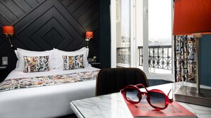 A hotel room with a bed, a nightstand, a table with a book and glasses, and a view of the city through large windows. - Hotel Des Arts Paris Montmartre (Paris)