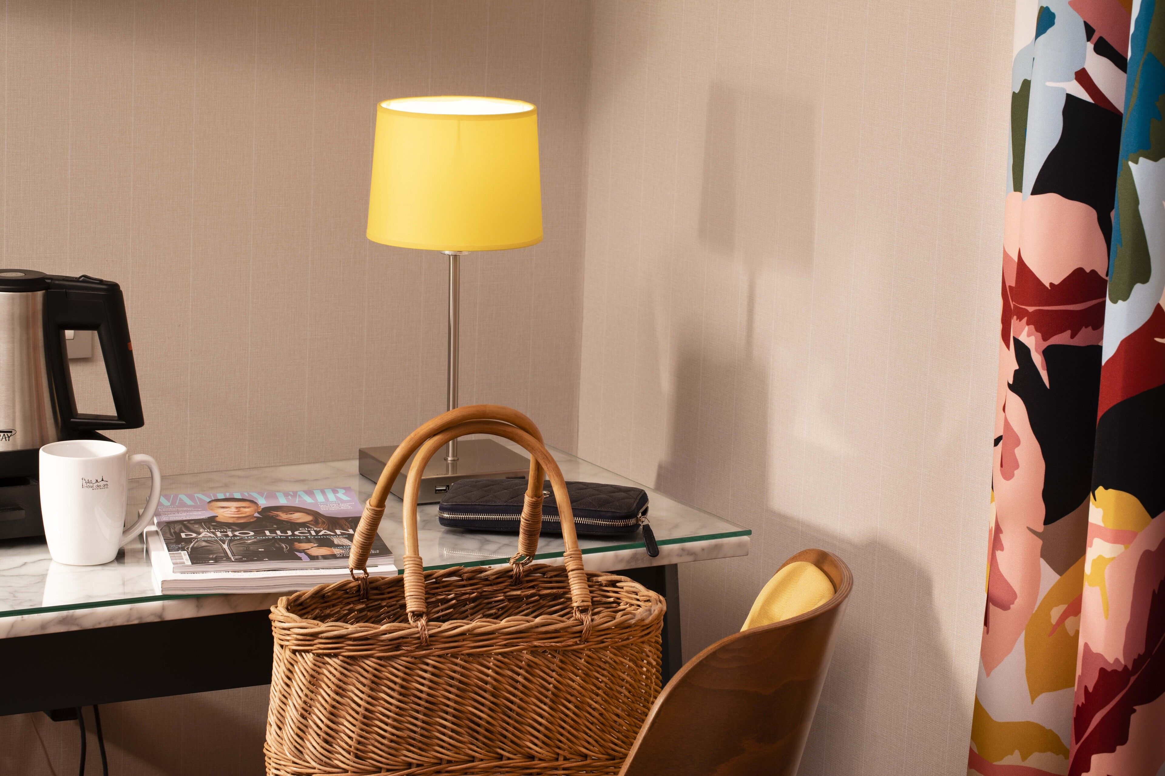 A hotel room with a glass-top desk, a wicker basket, a white mug, a magazine, a lamp, and a colorful curtain.